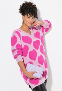 Overmaatse roze trui met grote donkerroze hartpatronen, v-halsontwerp en lange mouwen; gecombineerd met een lavendelkleurige gequiltte clutch.