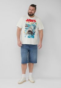 Weißes Baumwoll-T-Shirt mit einer bunten Grafik eines Hais und Vintage-Elementen. Getragen mit blauen Denim-Shorts und weißen Sneakers.