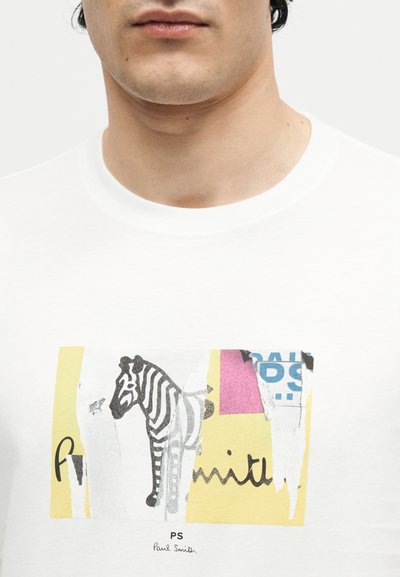 Κοντινό πλάνο ατόμου που φοράει άσπρη μπλούζα με σκισμένο σχέδιο ζέβρας και πολύχρωμα αφηρημένα σχήματα.