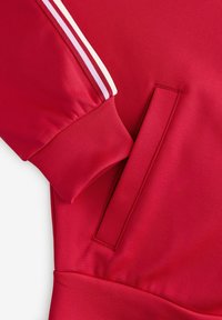 Manche de veste rouge avec des rayures blanches et roses près du poignet et une poche à fente verticale sur le devant.