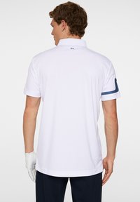 Vit kortärmad polo-shirt med en strukturerad krage, marinblå detaljer på ärmarna och en slät struktur. Har logotyp på nacken bak.