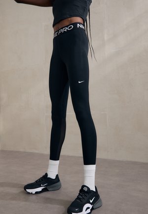 Personne portant des leggings Nike Pro noirs, des chaussettes blanches et des baskets Nike noires, se tenant contre un mur beige uni.