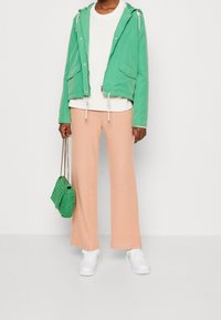 Zielona kurtka z kapturem na białym swetrze, zestawiona z brzoskwiniowymi szerokimi spodniami w prążki i białymi trampkami. Do stylizacji dołączona jest mała, zielona pleciona torba.