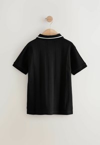 Polo nero esposto su un appendiabiti di legno, con maniche corte, colletto bianco a contrasto e texture a coste verticali.
