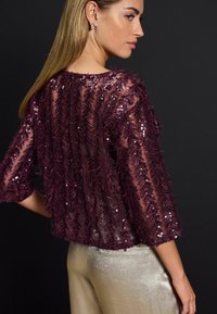 Top en sequins bourgogne avec manches trois-quarts, tissu transparent, motif de feuilles et texture légère, associée à un pantalon en satin couleur crème.