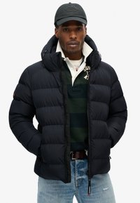 Sort puffersjakke med hætte, udstyret med lynlås, quiltet tekstur og frontlommer, båret over en mørkstribet polo skjorte og jeans.