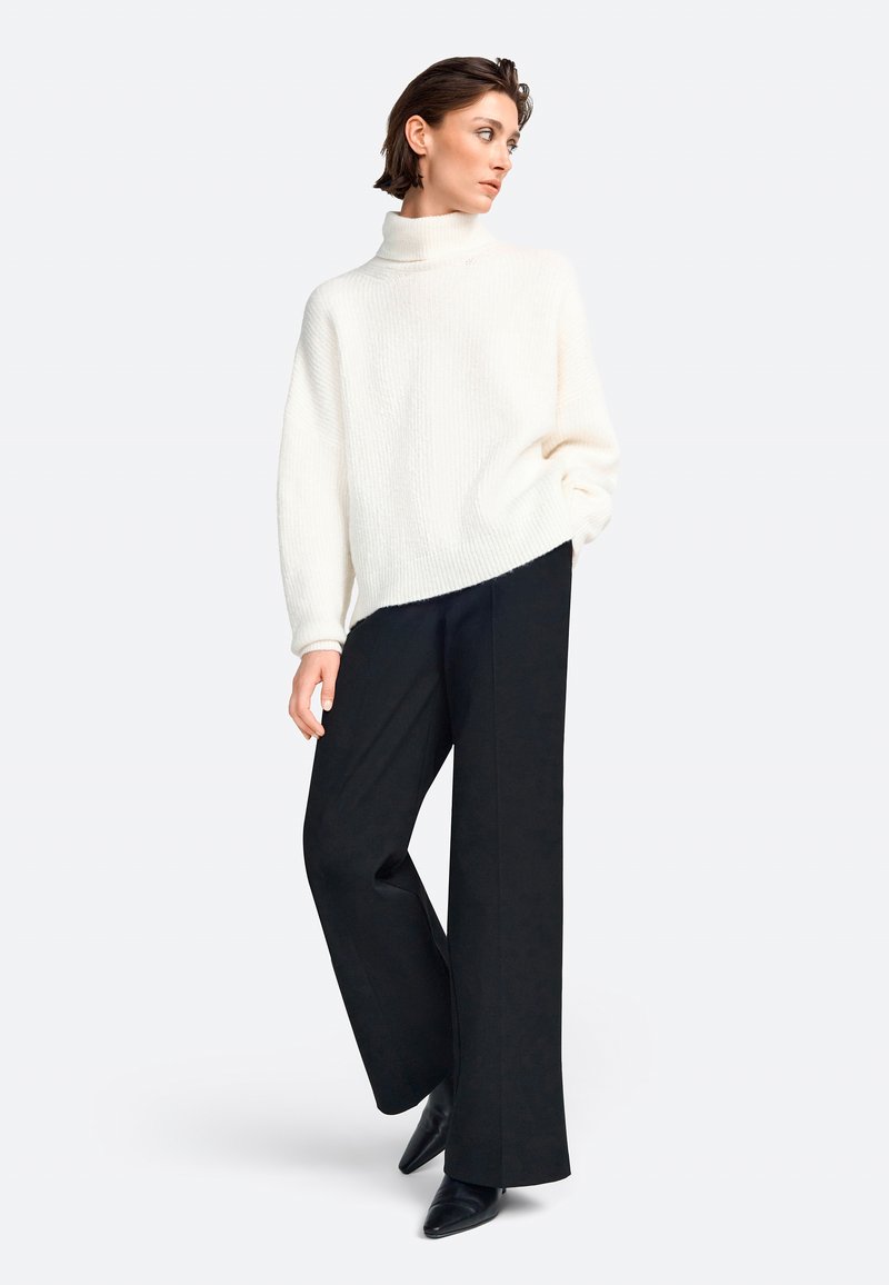 Weißer Strick-Turtleneck-Pullover kombiniert mit schwarzen weit geschnittenen Hosen und spitzen schwarzen Schuhen. Strukturiertes Material und lässige Passform.