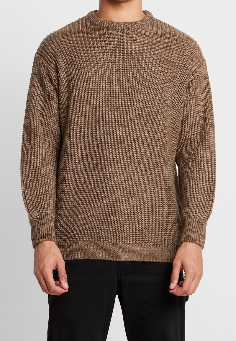 Person som bär en brun tjockstickad tröja med långa ärmar och ribbade mudd, i kombination med svarta byxor mot en enkel bakgrund.