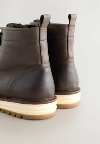 Par de botas de cuero marrón oscuro con suelas beige en capas, mostradas desde atrás sobre un fondo gris claro.