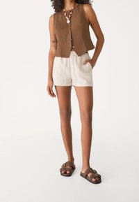 Brązowy bezrękawnik z guzikami, połączony z białymi szortami na sznurku i brązowymi klapkami, prezentujący swobodny letni strój.