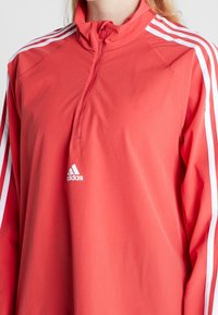 Punainen fleece-puloveri, jossa on puoliveto-aukko, ja hihassa valkoiset kolminkertaiset raidat sekä pieni valkoinen logo rinnassa.