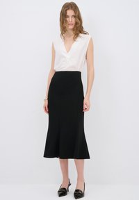 Valkoinen hihaton paita, jossa on V-kaula-aukko ja kultaiset yksityiskohdat, yhdistetty mustaan kellotettuun midi-hameeseen. Asusteena mustat kärjelliset korkokengät.