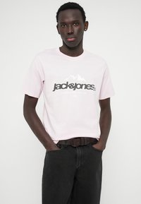 Ροζ βαμβακερό μπλουζάκι με εκτυπωμένο μαύρο λογότυπο και γραφικό βουνού, συνδυασμένο με σκούρες τζην παντελόνες και καφέ ζώνη.