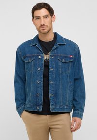 Jeansowa kurtka w ciemnoniebieskim kolorze z guzikami z przodu, dwoma kieszeniami na piersi i złożonym kołnierzem. Czarna koszulka widoczna pod nią. Luźny krój.