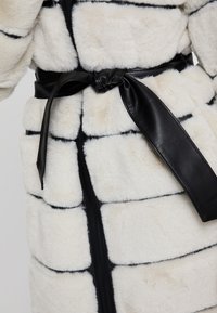 Bílý kabát z umělé kožešiny s černými pruhy, se černým koženým páskem a zipem vpředu. Měkká textura a nadýchané části.