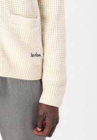 Suéter de punto color crema con un patrón texturizado, que presenta un bolsillo frontal y un detalle bordado en el dobladillo. Pantalones grises parcialmente visibles.