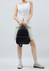 Musta nahkainen reppu, jossa on tekstuuripinta, etutasku ja kullatut yksityiskohdat. Malli pitää sitä käsissään, päällään valkoinen toppi ja kermaiset hame.