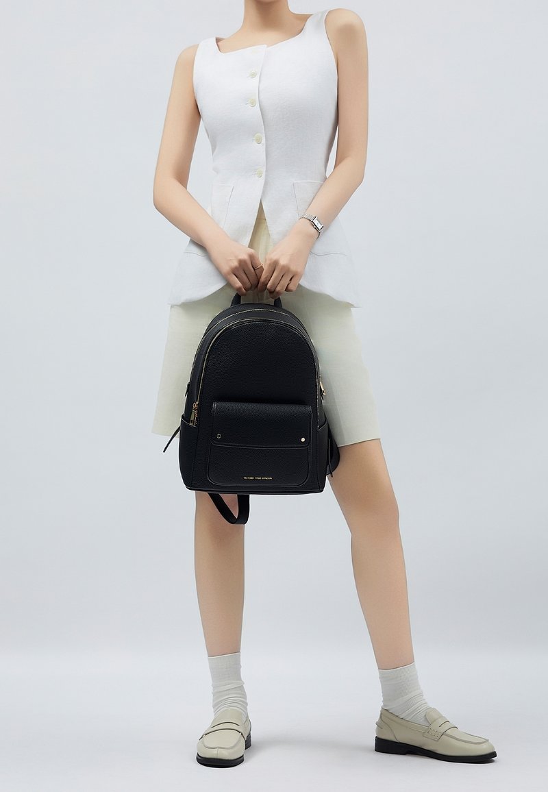 Musta nahkainen reppu, jossa on tekstuuripinta, etutasku ja kullatut yksityiskohdat. Malli pitää sitä käsissään, päällään valkoinen toppi ja kermaiset hame.