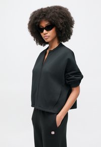 Czarna bluza na zamek o krótkim kroju, z prążkowanymi mankietami i zaokrąglonym kołnierzem, noszona z dopasowanymi czarnymi spodniami z małym logo.