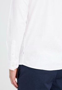 Camisa blanca de manga larga con puños abotonados, que presenta un dobladillo recto y un sutil detalle de etiqueta cerca de la costura lateral. Combinada con pantalones azul marino.