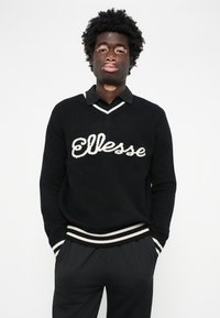 Sort sweater med hvid "Ellesse" logo, der har en v-hals design og stribede ribbede manchetter. Kombineret med sorte bukser for et sammenhængende look.