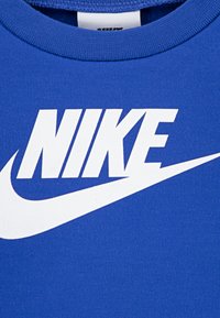 Tricou din bumbac albastru, cu un mare logo alb „NIKE” și design swoosh. Țesătura are o textură netedă și un guler cu margine canelată.