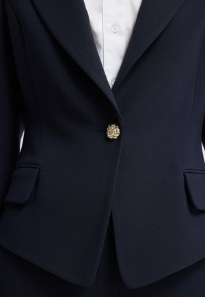 Blazer azul marino a medida con un diseño estructurado, que presenta un prominente botón en forma de cabeza de león dorado, dos bolsillos frontales y una tela texturizada.