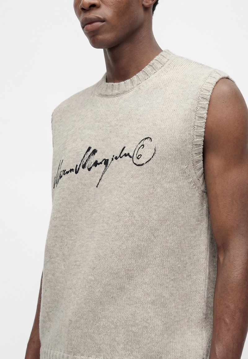 Mann trägt hellgrauen ärmellosen Strickpullover mit schwarzem kursivem Text auf der Brust vor einfarbig weißem Hintergrund.