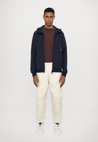 Jūrinės mėlynos zip-up tipo džemperis su dviem priekinėmis kišenėmis, derinamas su rudais marškiniais ir kreminės spalvos kelnėmis, apsirengtas su baltais sportbačiais.