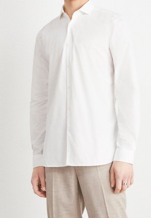 Mand iført en ensfarvet hvid langærmet skjorte med knapper og lysebeige bukser, stående mod en hvid baggrund.