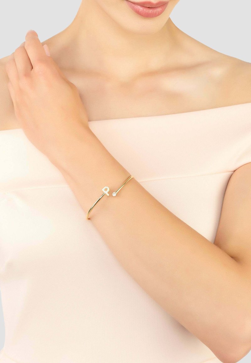 Arany karkötő, sima, vékony szalaggal, amelyen egy kis "R" betű és egy áttetsző drágakő dísz található, üres csuklón viselve.