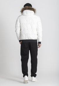 Giacca a vento bianca con cappuccio bordato di pelliccia, design trapuntato e polsini a coste neri, abbinata a pantaloni neri e sneaker bianche.
