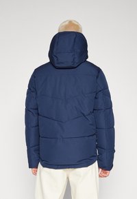 Giacca a vento blu navy con cappuccio, caratterizzata da pannelli trapuntati e polsini elasticizzati. La vista dall'indietro mostra una texture liscia e un design robusto.
