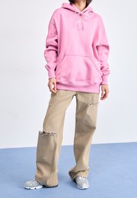 Person som bär en överdimensionerad rosa hoodie, beige rivna vida jeans och grå sneakers, står på blått golv mot en vit vägg.