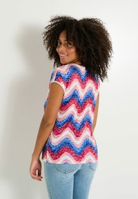 Top de manga corta con un estampado ondulado en rosa, rojo y azul, fabricado en una tela suave; presenta un cuello redondo y ligeras aberturas laterales.
