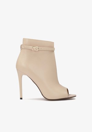 Beżowy botek na wysokim obcasie wykonany z gładkiej skóry, z odkrytymi palcami, cienkim paskiem z klamrą oraz smukłą sylwetką.