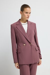 Blazer croisé en tissu mauve avec un col �à revers cranté, des boutons dorés et des poches avant. Porté sur un haut blanc avec un col décoratif.