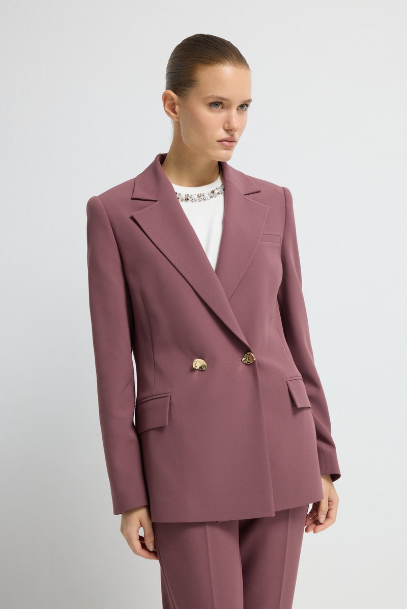 Blazer croisé en tissu mauve avec un col à revers cranté, des boutons dorés et des poches avant. Porté sur un haut blanc avec un col décoratif.