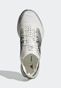 Zapatilla deportiva blanca con acentos de patrones negros, parte superior de textil, punta redonda, cierre de cordones y suela plana. Etiquetas visibles en el interior de la zapatilla.