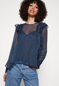 Blusa transparente azul marino con acentos de volantes, patrones florales en rosa que contrastan, mangas largas y un escote redondeado.