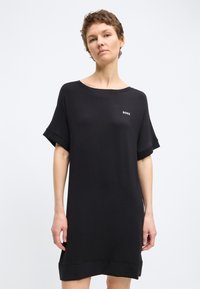 Γυναίκα που φοράει ένα φαρδύ μαύρο φόρεμα με κοντά μανίκια και μικρό λευκό λογότυπο "BOSS" στο στήθος, να στέκεται μπροστά σε μονόχρωμο φόντο.