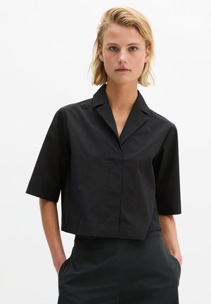 Femeie cu păr blond scurt, purtând o cămașă neagră largă cu mâneci scurte și guler și pantaloni negri, cu mâinile în buzunare, pe un fundal alb.