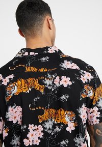 Zwart shirt met korte mouwen, versierd met levendige oranje tijgers en roze bloemmotieven, met een ontspannen kraag en zachte textuur.