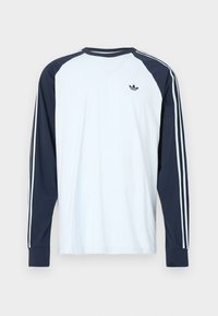 Λευκό μακρυμάνικο T-shirt με ναυτικό μπλε ραγκλάν μανίκια. Διαθέτει τρεις στενές λευκές ρίγες κατά μήκος των μανικιών και ένα μικρό μαύρο λογότυπο στο στήθος.