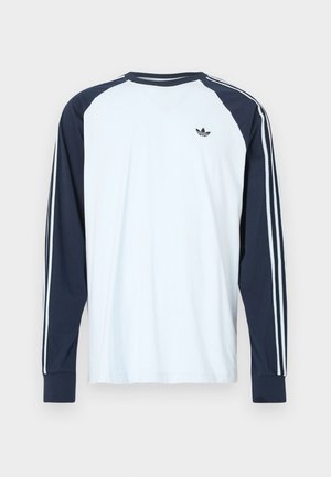 Λευκό μακρυμάνικο T-shirt με ναυτικό μπλε ραγκλάν μανίκια. Διαθέτει τρεις στενές λευκές ρίγες κατά μήκος των μανικιών και ένα μικρό μαύρο λογότυπο στο στήθος.