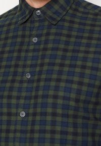 Zielono-granatowa koszula w kratę z kołnierzykiem na guziki, z ciemnymi guzikami i miękką, teksturowaną tkaniną. Widoczne detale wzoru w kratę.