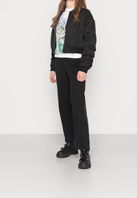 Černá bomber bunda s žebrovanými manžetami, bílý tričko s potiskem obsahující květinové ilustrace, a černé široké kalhoty doplněné chunky černými botami.