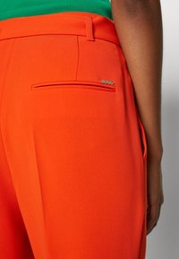 Gros plan sur une personne portant un pantalon ajusté orange vif avec une poche passepoilée à l'arrière et un petit logo métallique "BOSS".