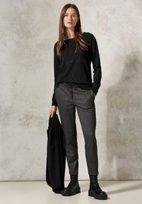 Maglione nero con motivo geometrico, pantaloni grigi texture e stivali neri con lacci. Il modello tiene in mano una giacca nera.