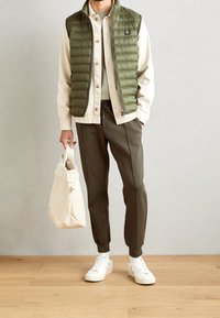 Olivgrön puffärmad väst över en beige skjorta, ihop med olivfärgade joggers och vita sneakers. Beige tote-väska med en knappdetalj.